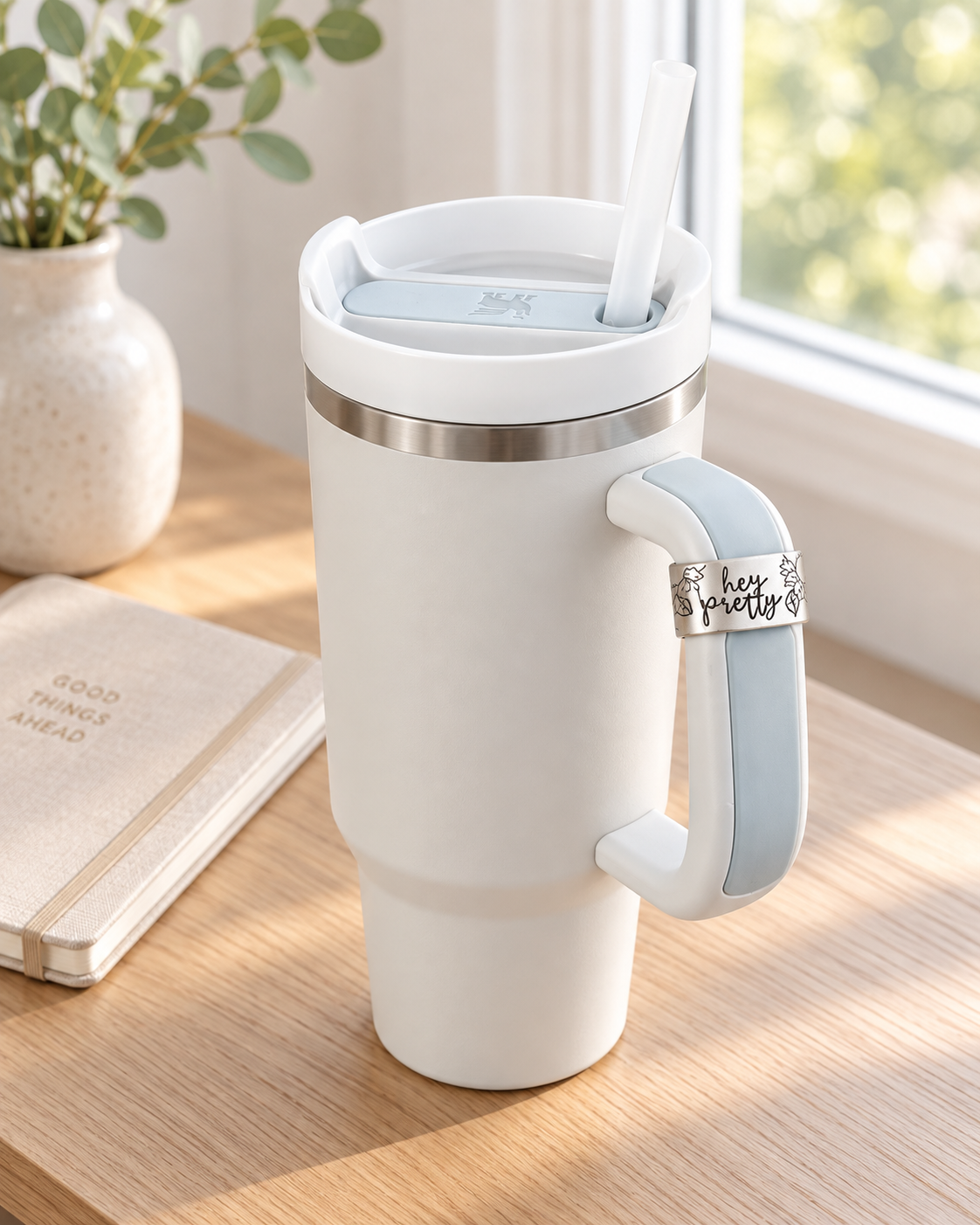 White handled tumbler with silver “Hey Pretty” floral stamped cuff displayed on a light wood desk.