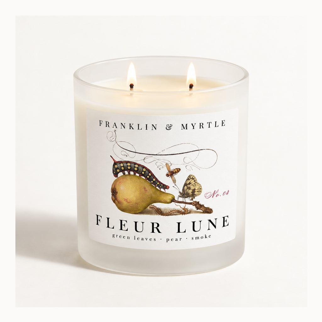 Fleur Lune double-wick candle burning in a frosted white glass jar with pear-and-butterfly label artwork.