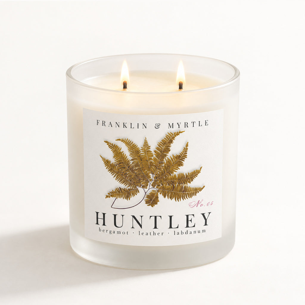 Huntley double-wick candle burning in a frosted white glass jar with golden fern label artwork.