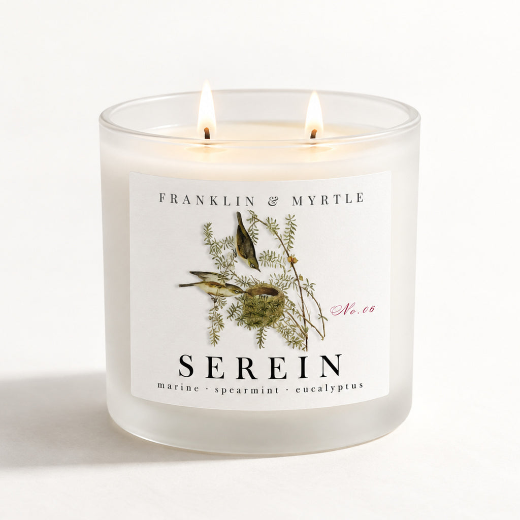 Serein double-wick candle burning in a frosted white glass jar with botanical bird label artwork.