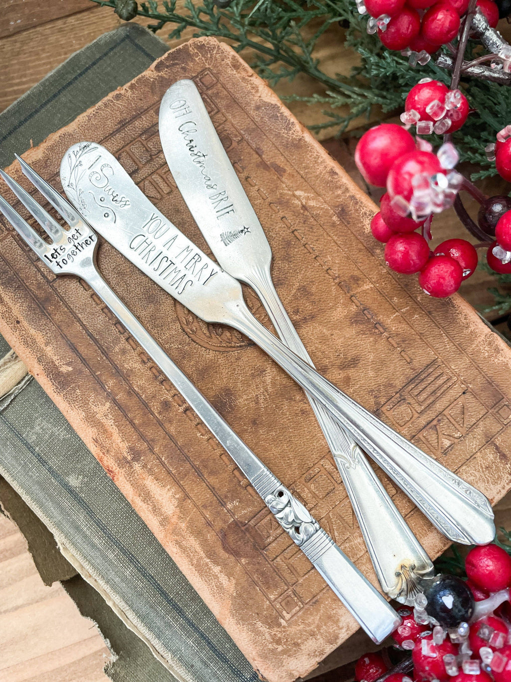 Vintage holiday charcuterie set with two hand-stamped spreaders and a cocktail fork styled with festive red berries on a rustic wooden board.