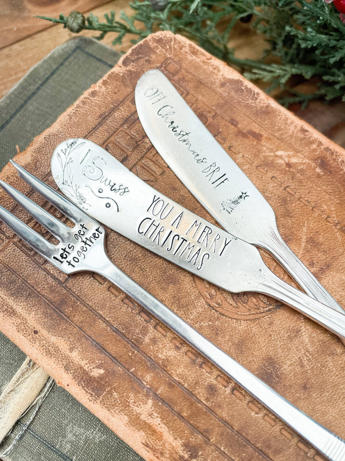 Vintage holiday charcuterie set with two hand-stamped spreaders and a cocktail fork arranged on a rustic wooden board.