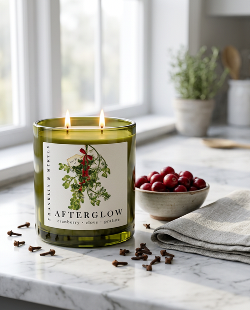 Lit Afterglow candle in an olive green glass jar on a marble counter beside a bowl of cranberries and scattered cloves.