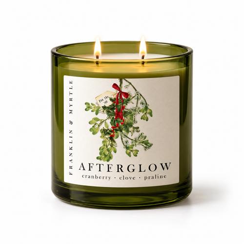 Lit Afterglow candle with two burning wicks in an olive green glass jar. Its label features mistletoe tied with a red ribbon and reads cranberry · clove · praline.
