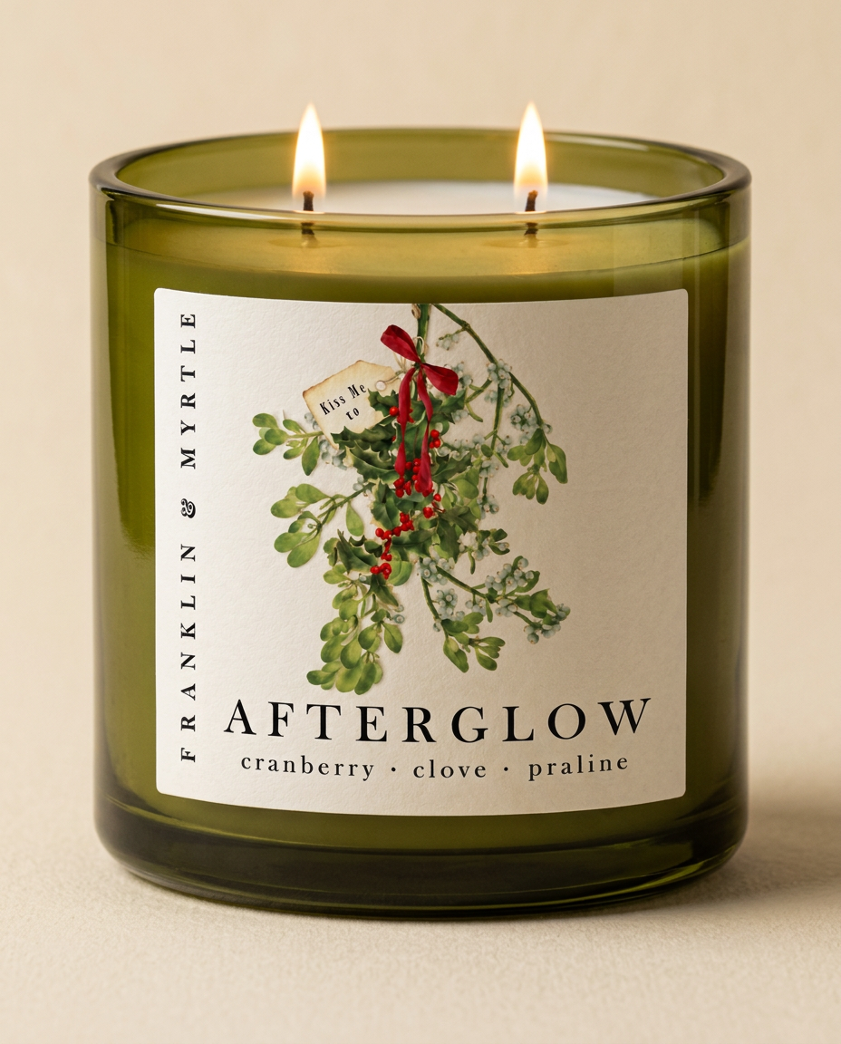 Lit Afterglow candle with two burning wicks in an olive green glass jar. Its label features mistletoe tied with a red ribbon and reads “cranberry · clove · praline.
