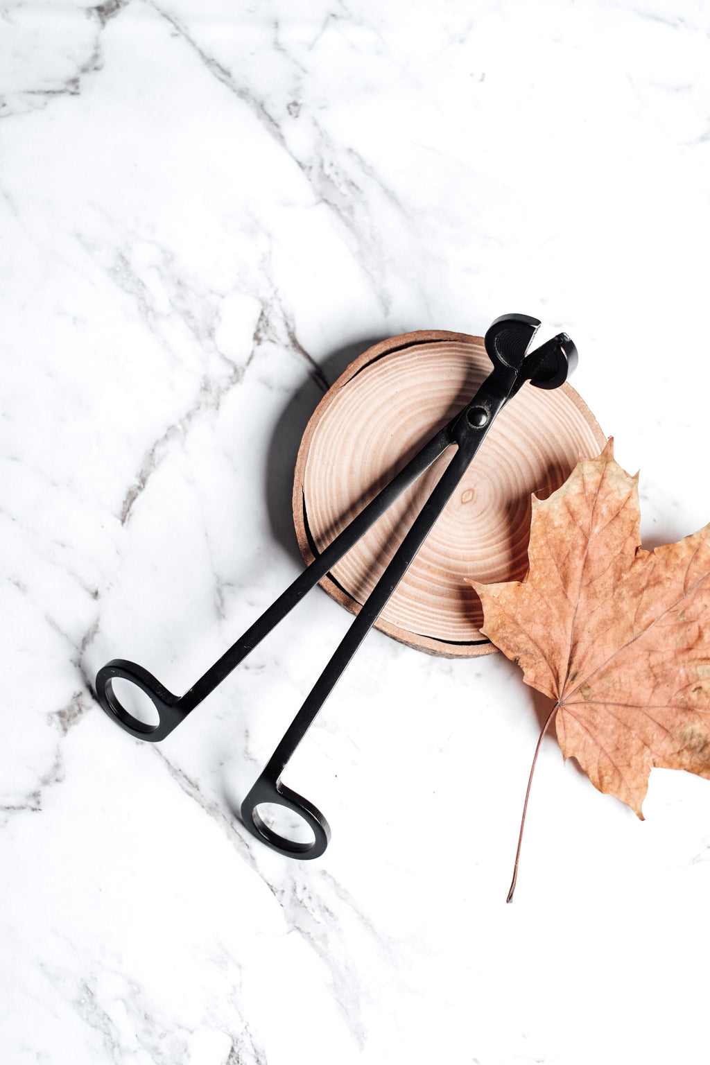 Black candle wick trimmer styled on a natural wood slice with a dried autumn leaf on a white marble background.