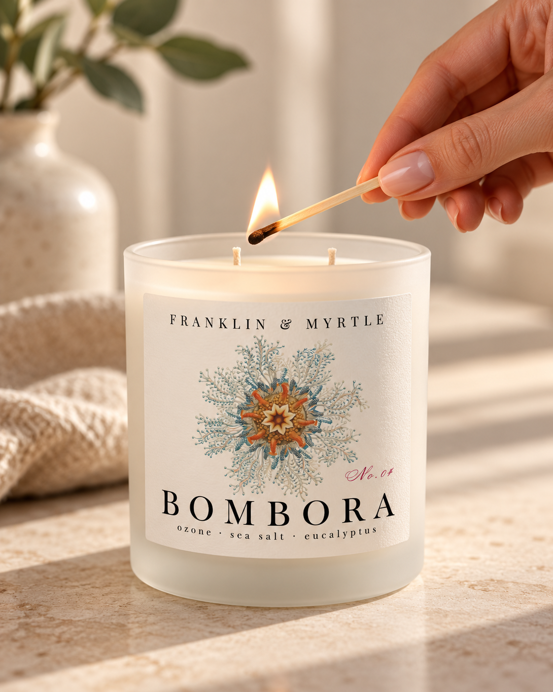 Hand holding a lit match above the two unlit wicks of a Bombora candle in frosted white glass.