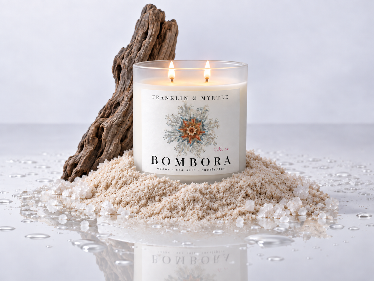 Lit Bombora double-wick candle in frosted white glass set in sea salt beside driftwood and water droplets.