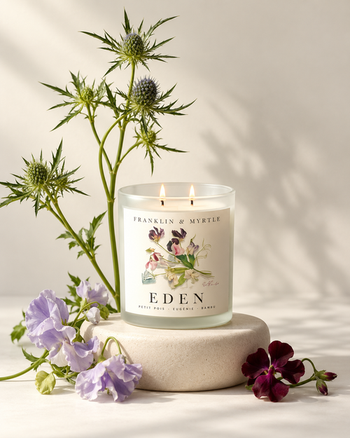 Lit Eden double-wick candle in frosted white glass on a stone pedestal surrounded by purple and burgundy flowers.