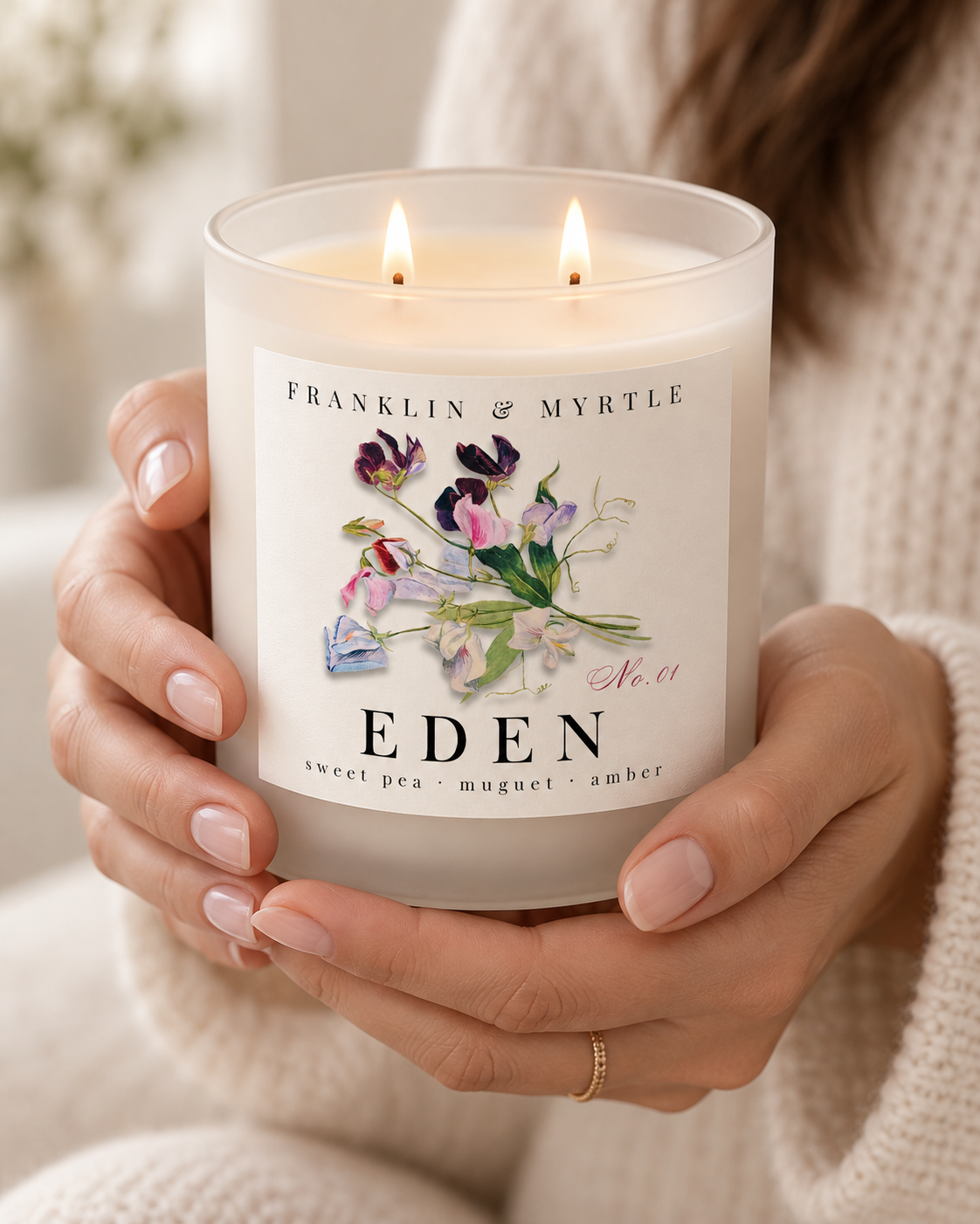 Person holding a lit Eden double-wick candle in a frosted white glass jar.