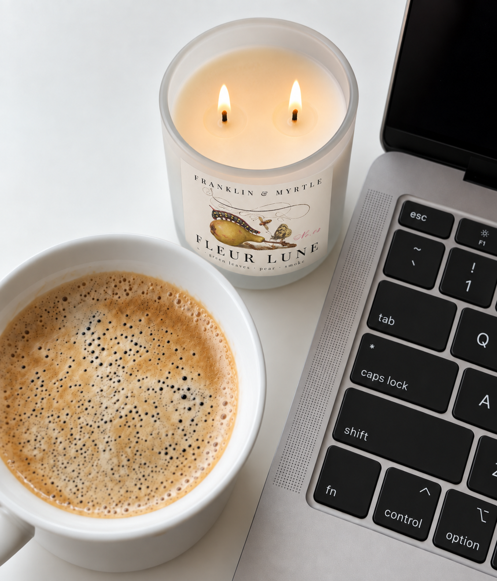 Lit Fleur Lune double-wick candle beside a cup of coffee and an open laptop on a white desk.