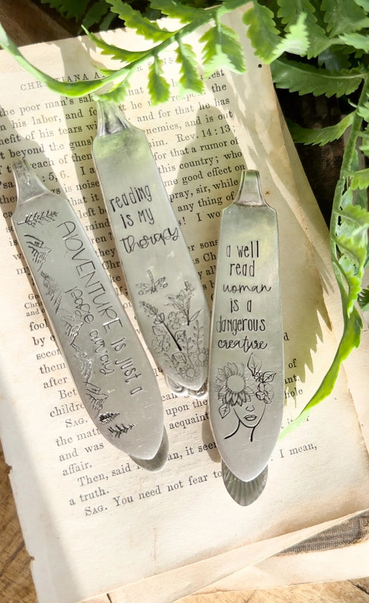 Three engraved metal reading bookmarks displayed on an open book with green fern leaves.