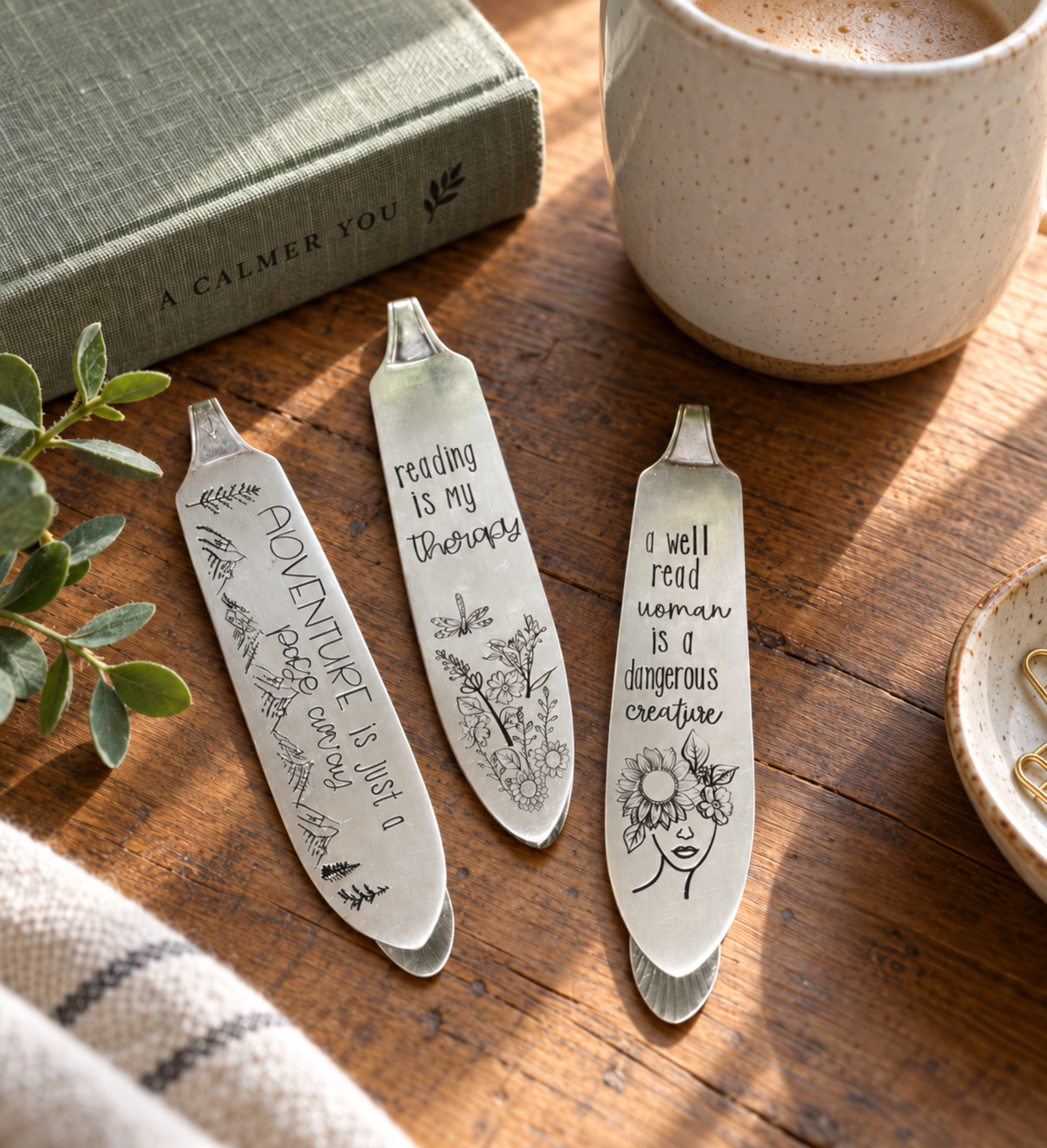 Three engraved metal reading bookmarks styled on a wooden table with a book, coffee mug, and greenery.