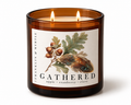 Lit Gathered candle with two burning wicks in an amber glass jar. Its label features oak leaves, acorns, and a feather above the notes apple, cranberry, and clove.