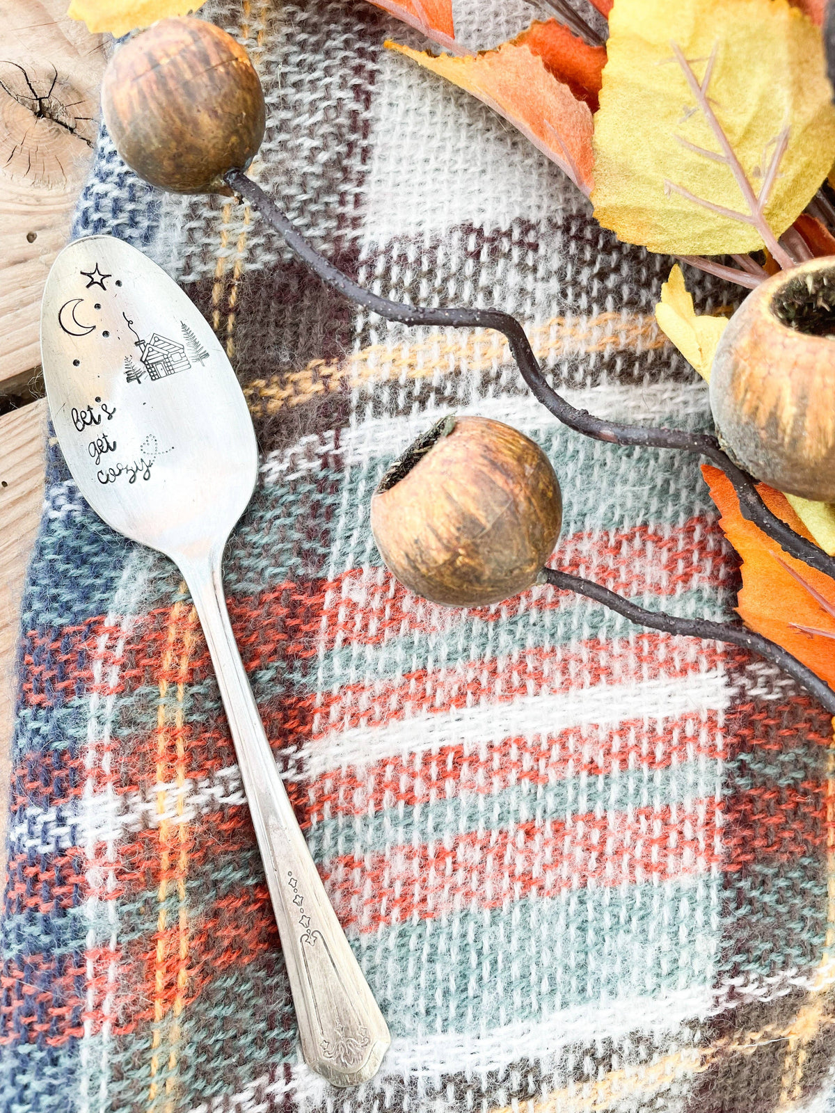 “Let’s Get Cozy” vintage spoon arranged on a plaid blanket with autumn leaves and acorn decorations.
