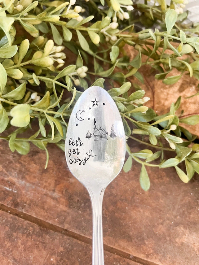 Silver-tone vintage spoon featuring “Let’s Get Cozy,” tiny cottages, trees, a crescent moon, and stars on a wooden surface with green foliage.
