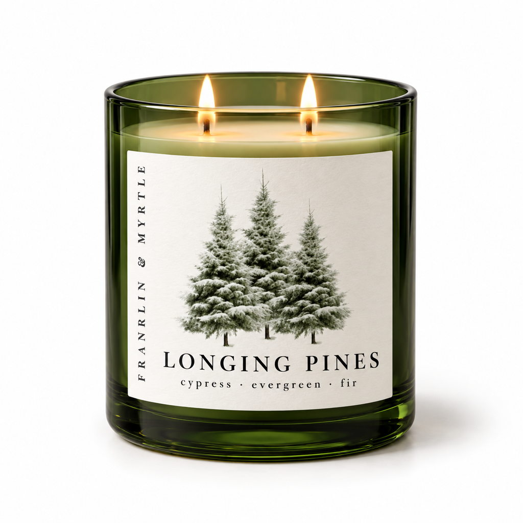 Longing Pines candle in a dark green glass jar with two lit wicks and a pine tree label against a light background.