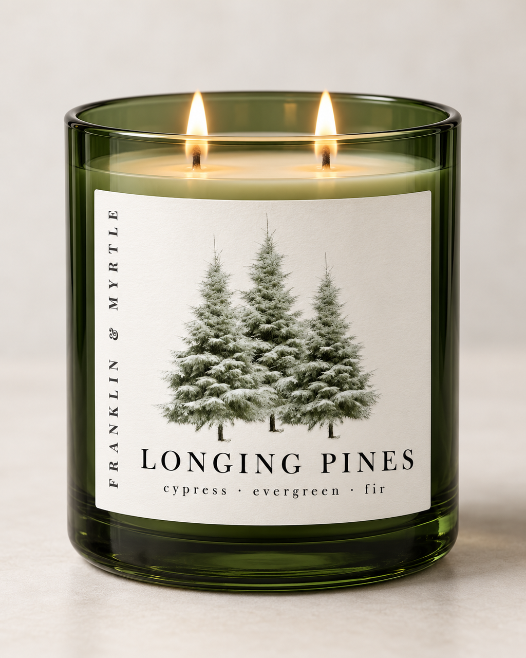 Longing Pines candle in a dark green glass jar with two lit wicks and a pine tree label against a light background.
