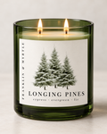 Longing Pines candle in a dark green glass jar with two lit wicks and a pine tree label against a light background.