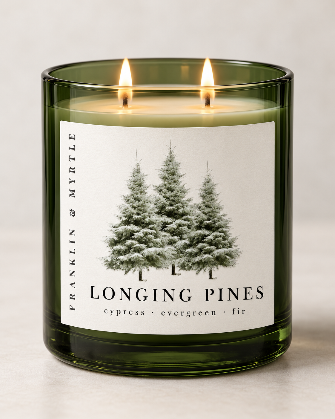 Longing Pines candle in a dark green glass jar with two lit wicks and a pine tree label against a light background.