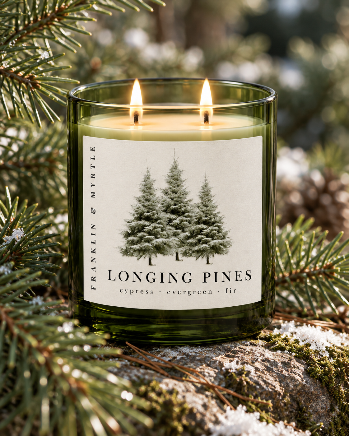 Lit Longing Pines candle in a green glass jar among evergreen branches on a mossy, lightly snow-dusted rock.