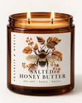 Lit Salted Honey Butter candle in an amber glass jar with two wicks and a label showing a bumblebee among autumn leaves