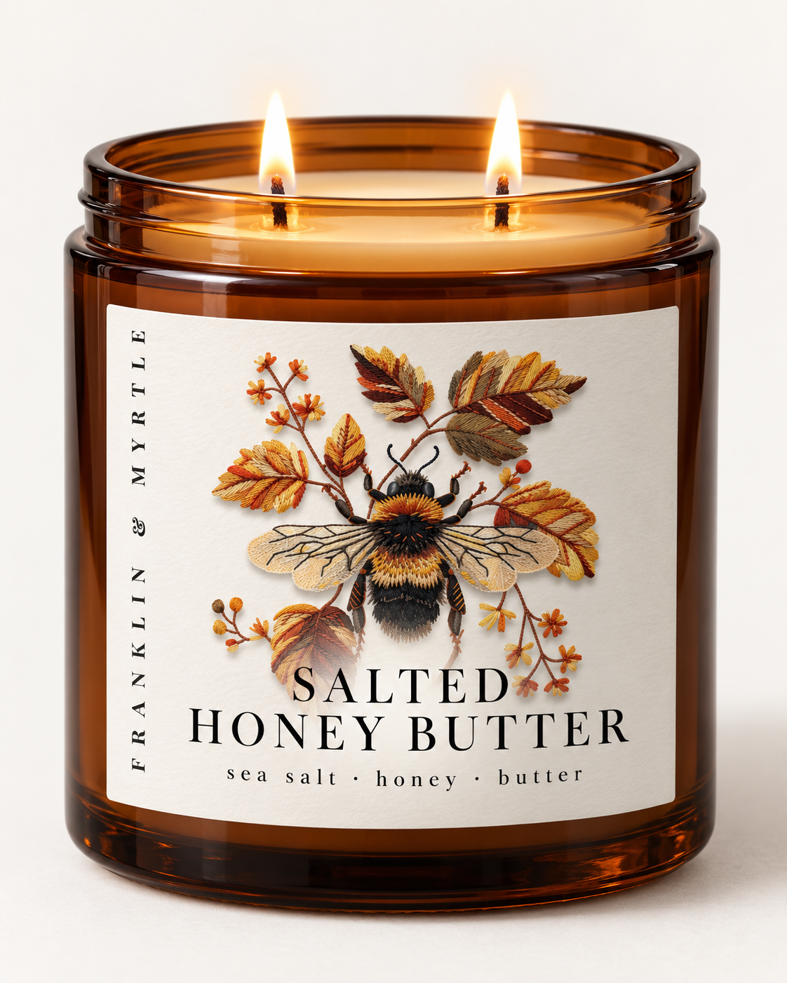 Lit Salted Honey Butter candle in an amber glass jar with two wicks and a label showing a bumblebee among autumn leaves