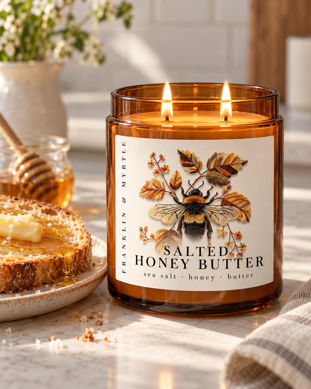 Lit Salted Honey Butter candle in an amber glass jar beside buttered toast drizzled with honey and a small jar of honey.