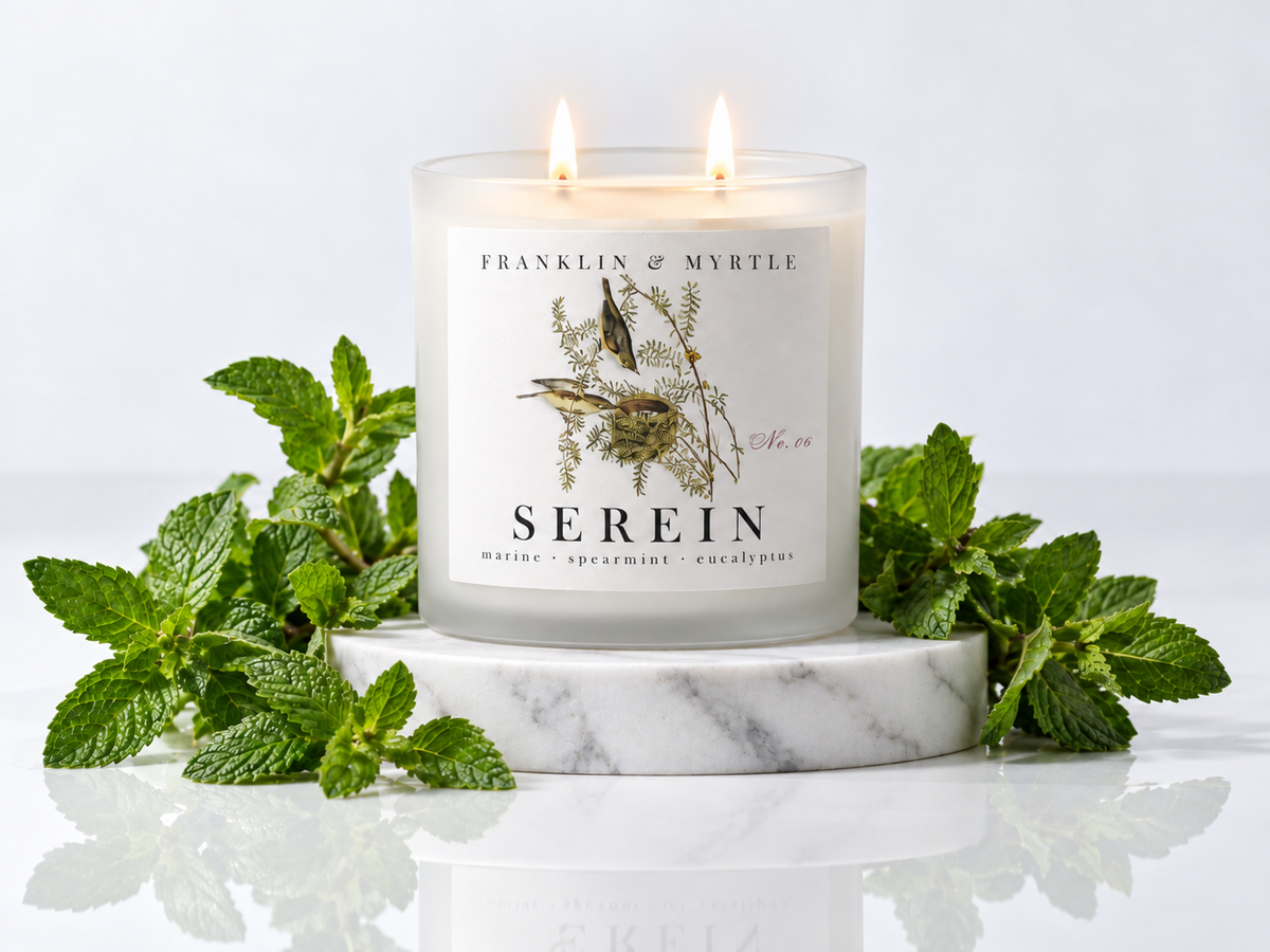 Lit Serein double-wick candle in frosted white glass on a marble pedestal surrounded by fresh mint.