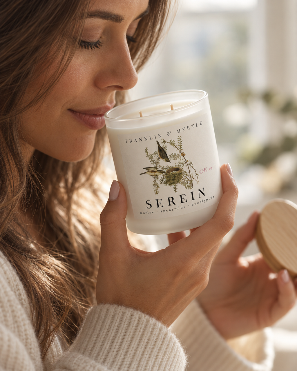Woman smelling an unlit Serein candle in a frosted white glass jar while holding its bamboo lid.