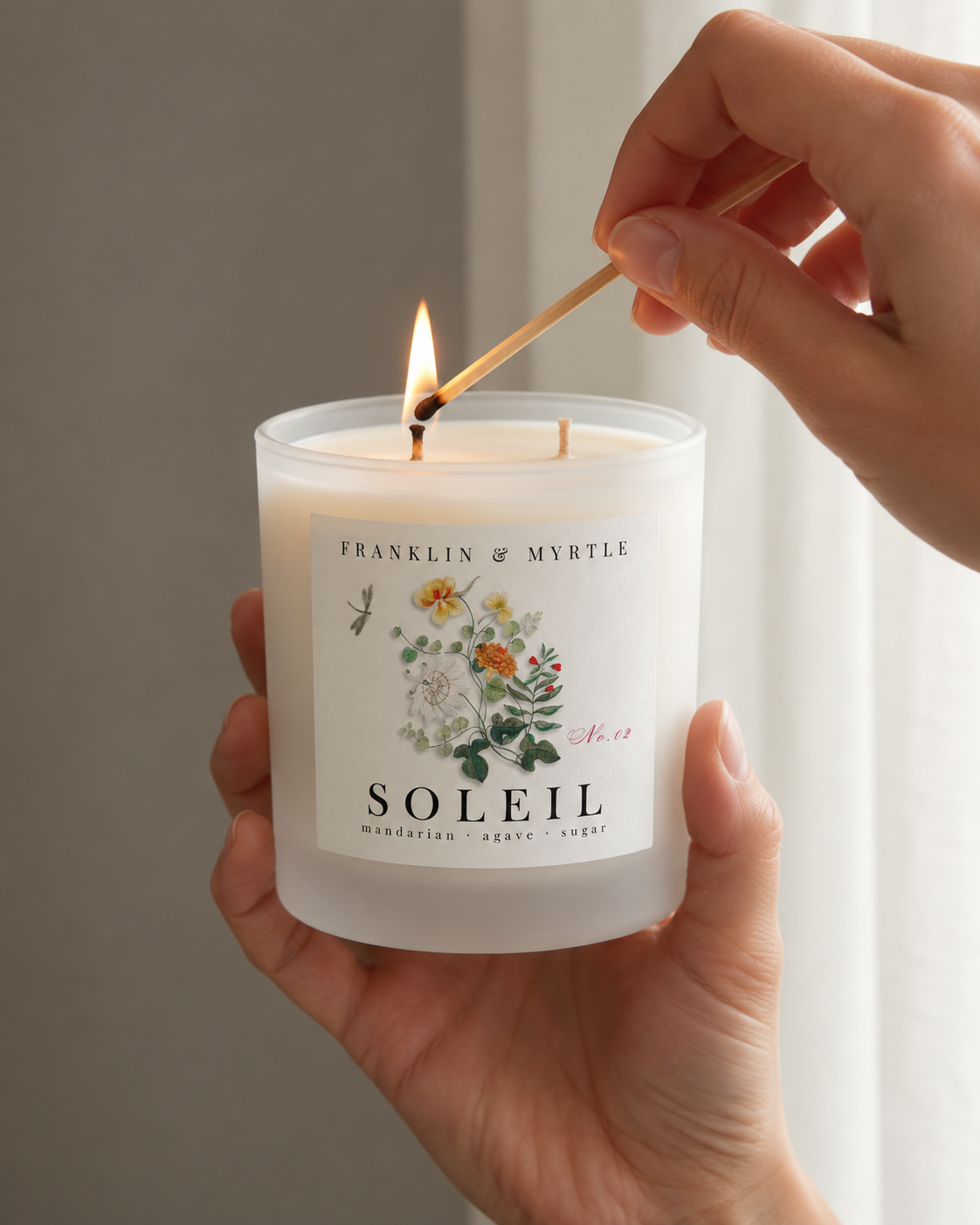 Hands hold a frosted white Soleil candle while a lit match lights one of its two wicks.