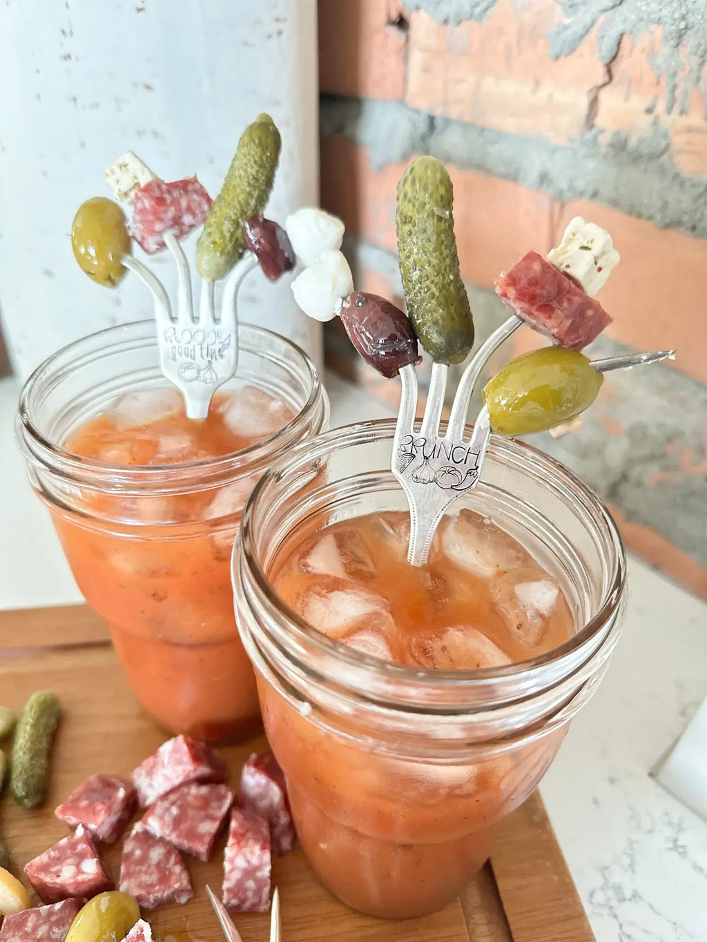 Two vintage hand-stamped Bloody Mary forks holding olives, pickles, cheese, and salami over Bloody Mary cocktails.