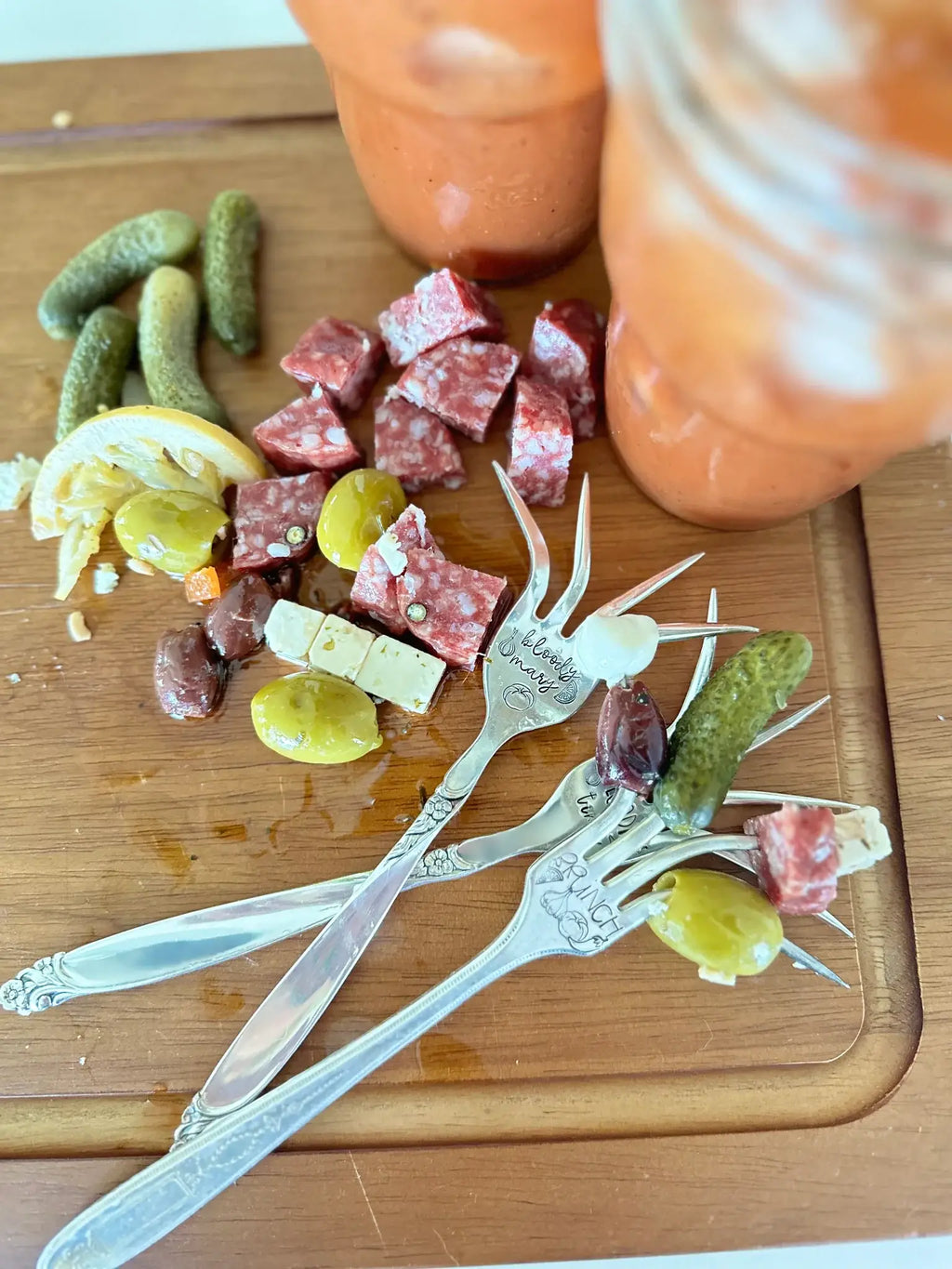 Two vintage hand-stamped Bloody Mary forks arranged with olives, pickles, salami, and cheese on a wooden board.