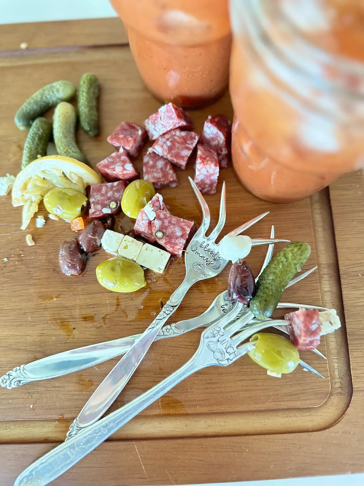 Two vintage hand-stamped Bloody Mary forks arranged with olives, pickles, salami, and cheese on a wooden board.