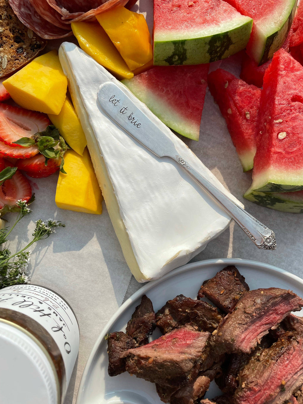 Vintage hand-stamped silver-plated spreader resting on brie beside fruit and sliced steak on a charcuterie board.