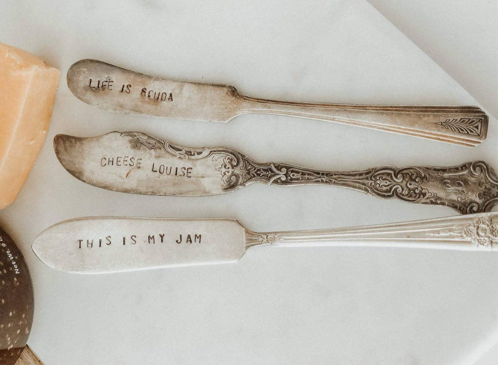 Three vintage silver-plated spreaders with hand-stamped sayings arranged on a white surface beside cheese.