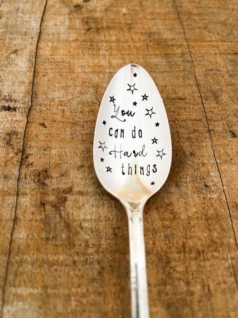 Vintage silver spoon stamped “You Can Do Hard Things” with star details on a rustic wood surface.